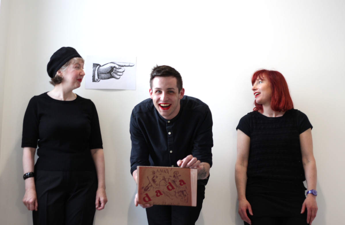 Portrait of three people wearing black and red lipkstick. Two white females face each other and one white male in the centre facing the camera and holding a picture with word Dada.