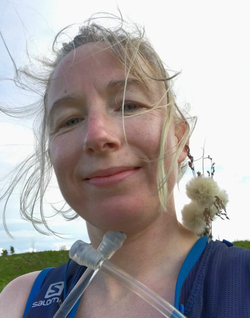 Head and shoulders shot of smiling female runner with wind in her tied-back blonde hair. She is wearing a running vest with water straws visible at the front, and she is carrying a posy of bog cotton in her backpack.